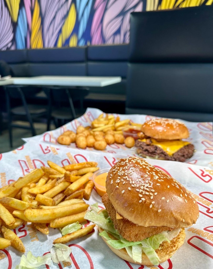 A vibrant table full of Craft Burger favourites at the Gosforth venue — featuring the Cheetos Smash Burger, New Yorker Boss Box, loaded chilli fries, Tayta tots, buffalo hot tenders, and house dips, all set against the fun, modern interior of the new branch.
