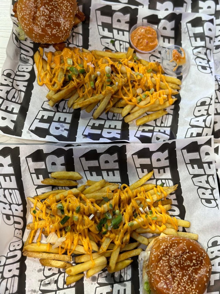 A vibrant table full of Craft Burger favourites at the Gosforth venue — featuring the Cheetos Smash Burger, New Yorker Boss Box, loaded chilli fries, Tayta tots, buffalo hot tenders, and house dips, all set against the fun, modern interior of the new branch.