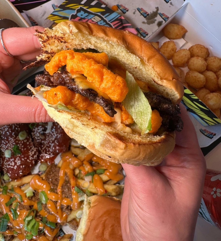Cheetos Smash Burger held above the table with loaded fries, buffalo tenders, and sides visible below.