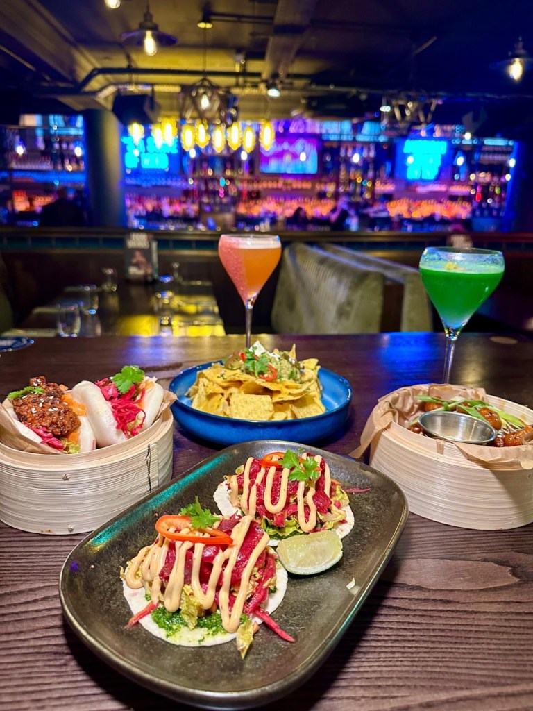 A table at Manahatta Newcastle featuring a mix of small plates and colourful cocktails, including frickles, salt and pepper squid, sticky chicken bites, waffle fries and vibrant signature drinks.