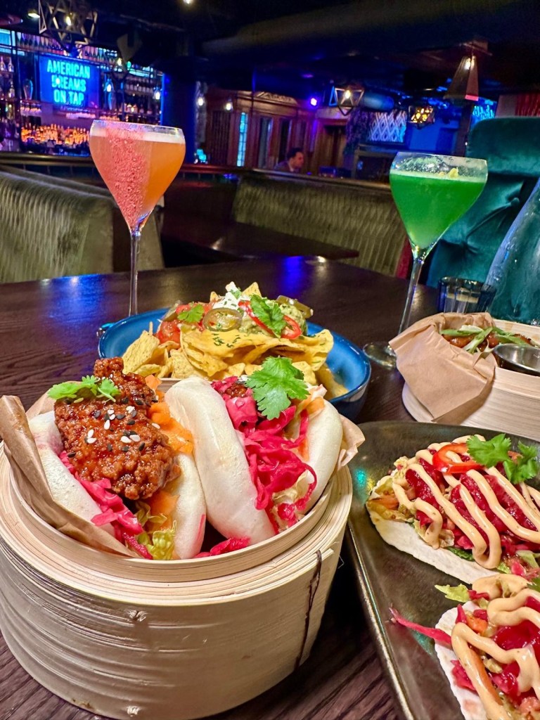 A table at Manahatta Newcastle featuring a mix of small plates and colourful cocktails, including frickles, salt and pepper squid, sticky chicken bites, waffle fries and vibrant signature drinks.